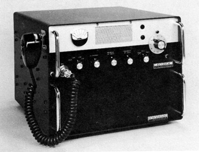Radio transceiver with 6 knobs, meter and speaker grill on front and microphone hung on left side.