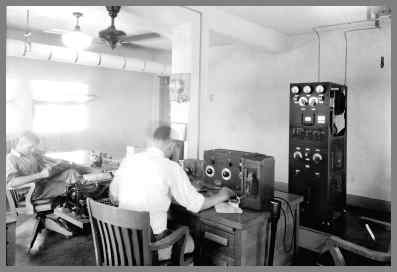 A later WUG2 radio room with transmitter in the background and 2 operators at desks in the foreground.