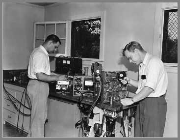 Two men working on two-way radio gear. Measurements Model 80 signal generator on the bench at the rear.