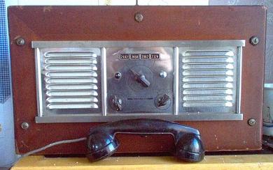 Tabletop radio transceiver with a channel selector knob in the center and a speaker grill both to the left and to the right of it.  2 smll knobs on lower front and a hsndset laying in front.
