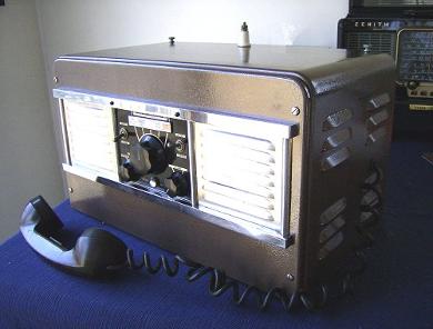 Tabletop radio transceiver with a channel selector knob in the center and a speaker grill both to the left and to the right of it.  2 smll knobs on lower front and a hsndset laying in front.