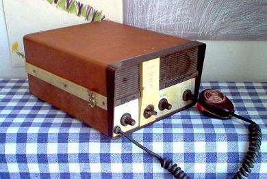Tabletop radio transceiver with a rectangular speaker grill on left front and 4 knobs below; microphone laying in front.