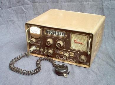 Tabletop Radio transceiver with a slide rule dial in the center, S meter on the left, square speaker grill on the right, 6 knobs and 3 toggle switches. Microphone laying in front.