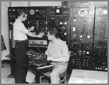 Five or six 6' racks of measurement gear in the background. One man making measurements with the other entering data on a tyoewriter.